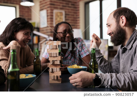 Laughing diverse people sitting at table while talking and enjoying society games together. Multiethnic people sitting at home in living room while discussing and playing with wood blocks. 90823057
