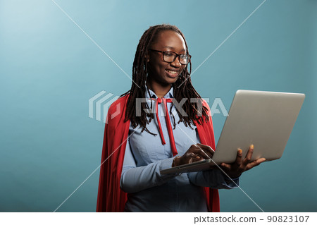 Brave and proud young adult superhero woman using modern laptop while standing on blue background. Confident and joyful justice defender wearing mighty hero cape while having handheld computer. Brave and proud young adult superhero woman using modern laptop while standing on blue background. Confident and joyful justice defender wearing mighty hero cape while having handheld computer. 90823107