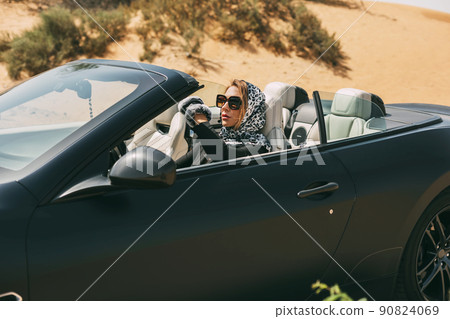 Glamorous blonde in a silk scarf and sunglasses in the driver's seat of a convertible convertible on a hot desert background. Fashion, style, glamour Glamorous blonde in a silk scarf and sunglasses in the driver's seat of a convertible convertible on a hot desert background. Fashion, style, glamour 90824069