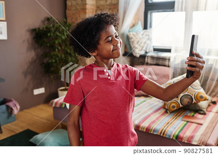 African smiling kid holding mobile phone in front of him and shooting video content standing in his room African smiling kid holding mobile phone in front of him and shooting video content standing in his room 90827581