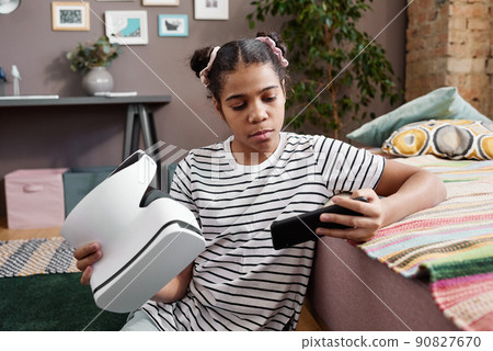 African girl sitting on floor in her room and trying to connect VR goggles with her smartphone to play game African girl sitting on floor in her room and trying to connect VR goggles with her smartphone to play game 90827670
