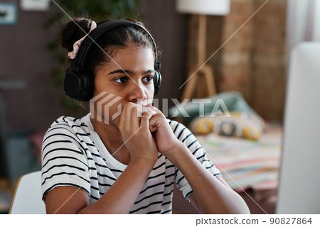 African serious girl in wireless headphones sitting in front of computer at home and looking at monitor 90827864