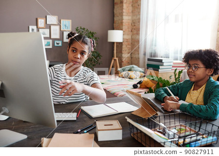 Girl pointing at computer monitor and discussing online presentation together with her little brother while they sitting at desk in room Girl pointing at computer monitor and discussing online presentation together with her little brother while they sitting at desk in room 90827911