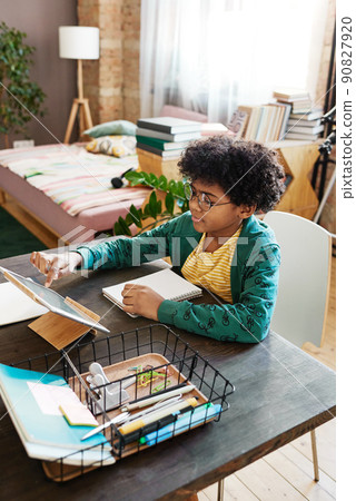 African little boy sitting at table with notebook and touching the screen of digital tablet, he studying at home African little boy sitting at table with notebook and touching the screen of digital tablet, he studying at home 90827920