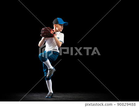 Dynamic portrait of little baseball player, pitcher in blue-white uniform training isolated on black studio background. Concept of sport, achievements, studying, competition 90829488