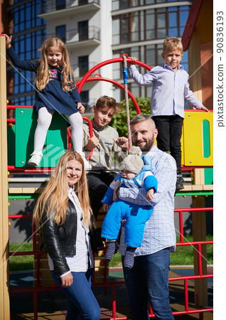 Happy family - father, mother and children having fun together on playground. Three cheerful kids playing while parents holding toddler in arms. Modern residential buildings on background. 90836193