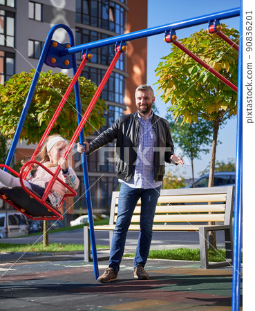 Bearded father carefully pushing swing with daughter, smiling at sunny morning. Handsome man swinging adorable daughter at modern courtyard of city residential high-rise buildings. 90836201