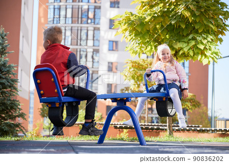 Cheerful brother and sister, in warm clothes, swinging at modern courtyard of city residential high-rise buildings.. Side view of two little friends having fun, sitting on see-saw swing at playground. Cheerful brother and sister, in warm clothes, swinging at modern courtyard of city residential high-rise buildings.. Side view of two little friends having fun, sitting on see-saw swing at playground. 90836202