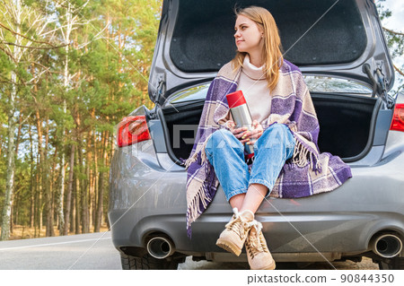 Young woman sitting on car trunk with coffee cups and thermos on the side of the road in the forest in warm fall or spring day. Time for yourself. Contemplation and rest in solitude. Young woman sitting on car trunk with coffee cups and thermos on the side of the road in the forest in warm fall or spring day. Time for yourself. Contemplation and rest in solitude. 90844350