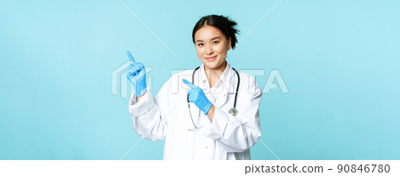 Smiling female doctor, physican in medical uniform and sterile gloves, pointing fingers left at promo, clinic logo, standing over blue background 90846780
