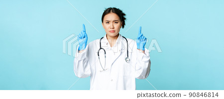 Angry and concerned female doctor, physician pointing fingers up, frowning disappointed, standing in medical uniform over blue background 90846814