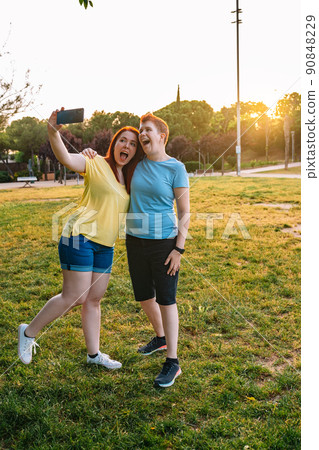 two smiling friends taking a selfie, in a public park in the city at sunset. young girl couple enjoying the summer outdoors.concept of friendship and companionship. vertical 90848229