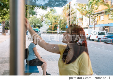 young girls waiting for the city bus at the bus stop. woman checking the bus route, looking at the information board. photograph through glass. 90848301