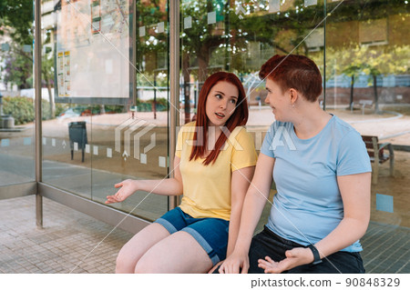 two happy young friends sitting at the bus stop, talking and gossiping. young girls in the city. concept of friendship and companionship. 90848329