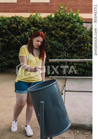young urban girl with red hair, picking up rubbish from the ground to put it in a bin. young environmentalist in the city. vertical. 90848351