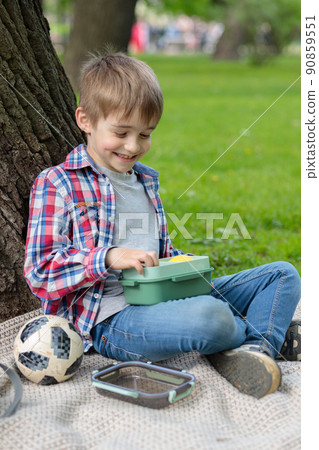 Lunch in the fresh air. A boy sitting by a tree, eating his lunch from a lunch box. Selective focus. 90859551
