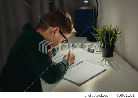 caucasian boy doing homework at home in dark room by the light of lamp. 90861191