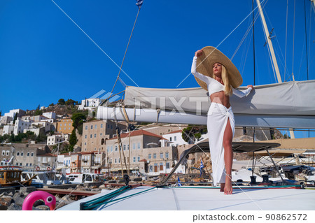 Beautiful young blond woman standing on catamaran at sunny summer day at Hydra marina bay, Greece Beautiful young blond woman standing on catamaran at sunny summer day at Hydra marina bay, Greece 90862572