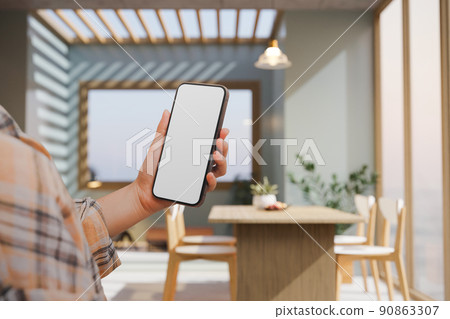 A female hand holding a smartphone mockup over blurred comfortable beautiful dining room 90863307