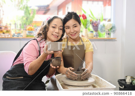 Smiling middle aged woman and daughter creating handmade tableware in pottery workshop. Pottery working, handmade clay products and creative skills 90866418
