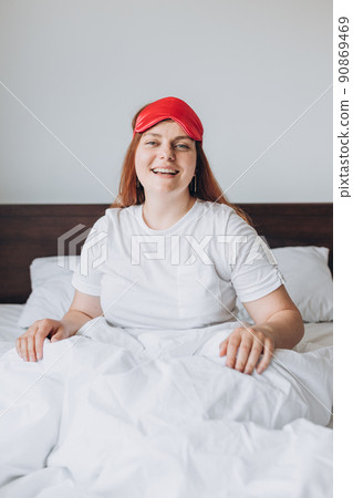 Cheerful young happy woman in pajama smiling and lifting sleep mask after awakening in morning. Redhead girl waking up enjoy good morning looking at camera 90869469