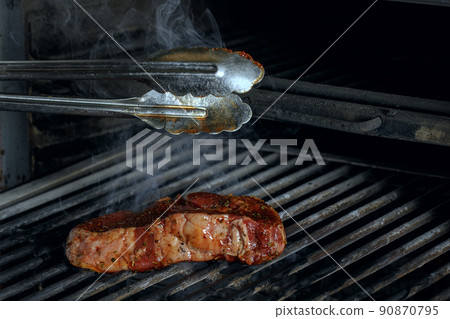 using silver serving kitchen tongs holding raw meat beef steak bbq grilling on rack charcoal stove with smoke on black background. 90870795