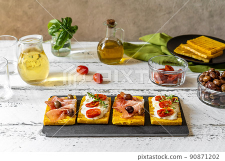 Grilled polenta with stracchino cheese, tomatoes, herbs, speck and olive. White wooden background, ingredients and table wine. Grilled polenta with stracchino cheese, tomatoes, herbs, speck and olive. White wooden background, ingredients and table wine. 90871202