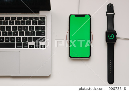 Laptop with smart watch and phone charging on wireless charger. Green screen on phone, on-screen charging indicator on watch. Top view. Place for text Laptop with smart watch and phone charging on wireless charger. Green screen on phone, on-screen charging indicator on watch. Top view. Place for text 90872848