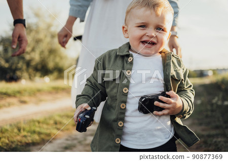 Funny Toddler Boy Holding Toys in Hands and Running Away from His Parents Outdoors 90877369