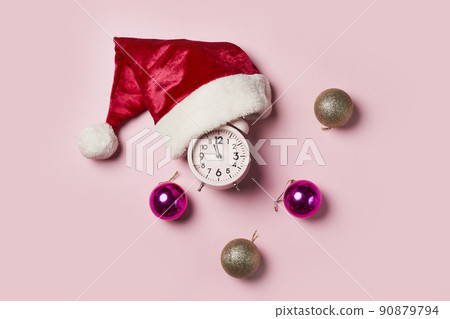 Christmas time consept.Christmas clock in a Santa Claus hat or cap on a pink background. The minute hand at twelve o'clock on New Year's Eve. Christmas time consept.Christmas clock in a Santa Claus hat or cap on a pink background. The minute hand at twelve o'clock on New Year's Eve. 90879794