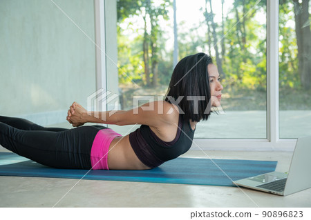 Sporty woman practicing yoga, back and buttock training pose,.stretching watching fitness video tutorial online on laptop, doing workout at home sitting on mat in living room practicing 90896823