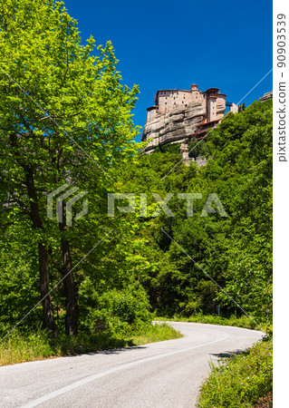A curved road with the Rusanu Monastery on a huge rocky mountain in Meteora, Greece 90903539