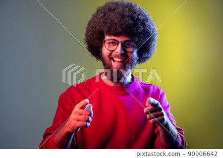 Portrait of hipster man with Afro hairstyle pointing to camera, smiling, making happy choice, we need you concept, wearing red sweatshirt. Indoor studio shot isolated on colorful neon light background 90906642