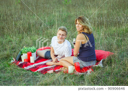 Beautiful young happy loving couple on picnic lying down on plaid in field on sunny summer day enjoying, holding and drinking wine and resting. looking at camera and smiling. 90906646