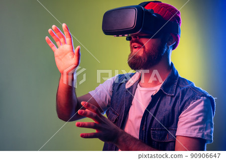 Impressed bearded hipster man standing in vr, playing video game and trying to touch something, wearing beanie hat and denim vest. Indoor studio shot isolated on colorful neon light background. 90906647
