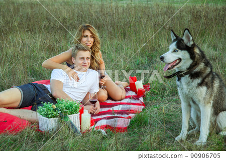 Beautiful young happy loving couple on picnic lying down on plaid with their dog in field on sunny summer day enjoying, holding and drinking wine and resting. looking at their dog and smiling. Beautiful young happy loving couple on picnic lying down on plaid with their dog in field on sunny summer day enjoying, holding and drinking wine and resting. looking at their dog and smiling. 90906705