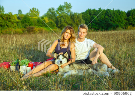 Beautiful young happy loving couple on picnic lying down on plaid with their dog in field on sunny summer day enjoying and resting. looking at camera and smiling. Beautiful young happy loving couple on picnic lying down on plaid with their dog in field on sunny summer day enjoying and resting. looking at camera and smiling. 90906962