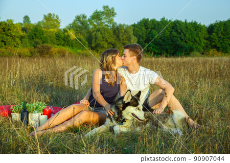 Beautiful young happy loving couple on picnic lying down on plaid with their dog in field on sunny summer day enjoying and resting. kissing and have fun. Beautiful young happy loving couple on picnic lying down on plaid with their dog in field on sunny summer day enjoying and resting. kissing and have fun. 90907044