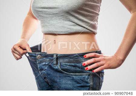 Closeup of slim waist of young woman standing in big jeans and gray top showing successful weight loss, indoor studio shot, isolated on light gray background, diet concept. 90907048