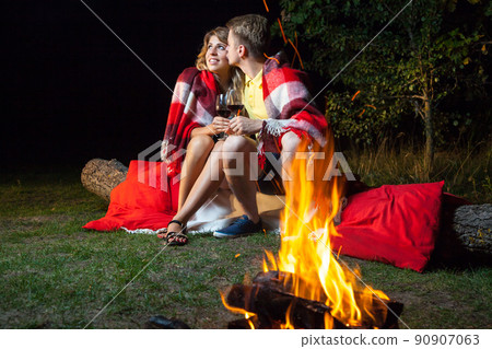 Young happy loving couple sitting on log near the fire covered with plaid holding wine, drinking and enjoy their romantic night. he is kissing her and smiling. 90907063