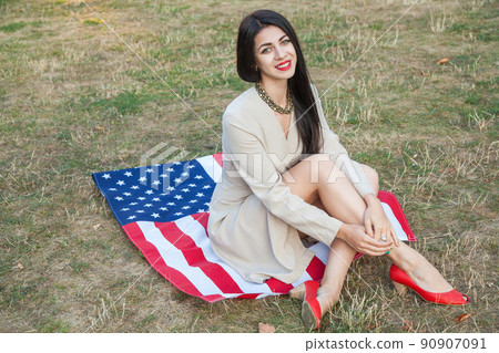 Beautiful sexy young woman with classic dress lying down on american flag in the park. fashion model holding us smiling and looking at camera. USA lifestyle with toothy smile. 90907091