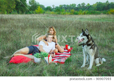 Beautiful young happy loving couple on picnic lying down on plaid with their dog in field on sunny summer day enjoying, holding and drinking wine and resting. looking at their dog and smiling. Beautiful young happy loving couple on picnic lying down on plaid with their dog in field on sunny summer day enjoying, holding and drinking wine and resting. looking at their dog and smiling. 90907093