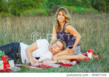 Beautiful young happy loving couple on picnic lying down on plaid in field on sunny summer day enjoying, holding and drinking wine and resting. looking at camera and smiling. Beautiful young happy loving couple on picnic lying down on plaid in field on sunny summer day enjoying, holding and drinking wine and resting. looking at camera and smiling. 90907109
