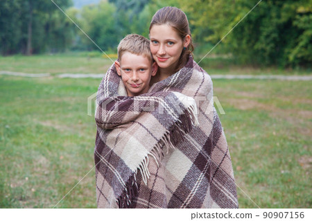 Young sister and brother with freckles on their faces stand covered with plaid in park, get warm, smiling and looking at camera. 90907156