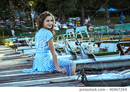 Beautiful young fashion model with freckles on her face and light blue striped dress and fashion makeup and hairstyle is sitting on pier near metalic silver boat on lake, posing and looking into the 90907269