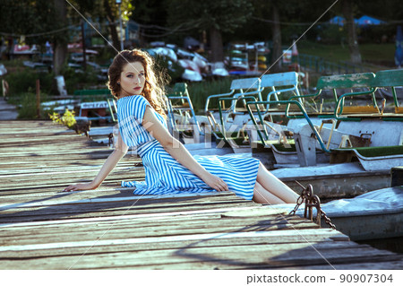 Beautiful young fashion model with freckles on her face and light blue striped dress and fashion makeup and hairstyle is sitting on pier near metalic silver boat on lake, posing and looking into the Beautiful young fashion model with freckles on her face and light blue striped dress and fashion makeup and hairstyle is sitting on pier near metalic silver boat on lake, posing and looking into the 90907304