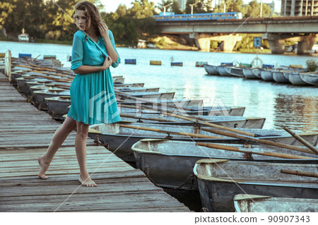 Beautiful young fashion model in turquois dress and fashion makeup and hairstyle is posing on pier, posing and looking into the distance on wood road. 90907343