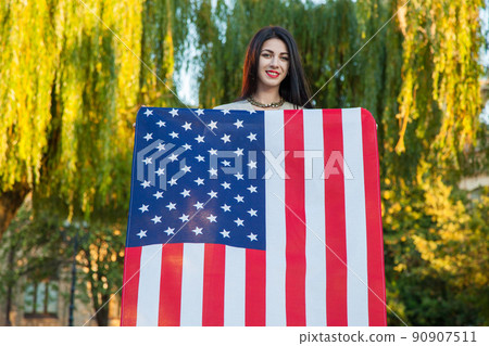 Beautiful young woman with classic dress holding american flag in the park. fashion model holding us smiling and looking at camera. usa lifestyle. 90907511