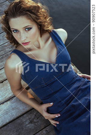 Beautiful young fashion model with freckles on her face and denim blue dress and fashion makeup and hairstyle is lying down on pier, posing and looking at camera. 90907523