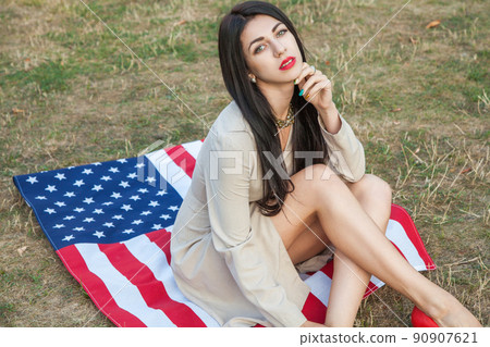 Beautiful sexy young woman with classic dress lying down on american flag in the park. fashion model holding us smiling and looking at camera. USA lifestyle with toothy smile. Beautiful sexy young woman with classic dress lying down on american flag in the park. fashion model holding us smiling and looking at camera. USA lifestyle with toothy smile. 90907621